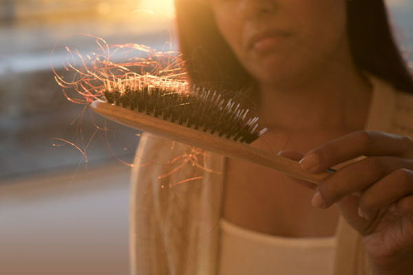 woman lit from behind has a concerned expression as she holds a hairbrush in front of her with a substantial amount of hair in it, suggesting hair loss